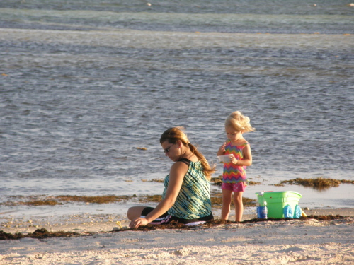 mother sitting facing the ocean sifting the beach sand and young daughter standing behind her throwing sand in the wind